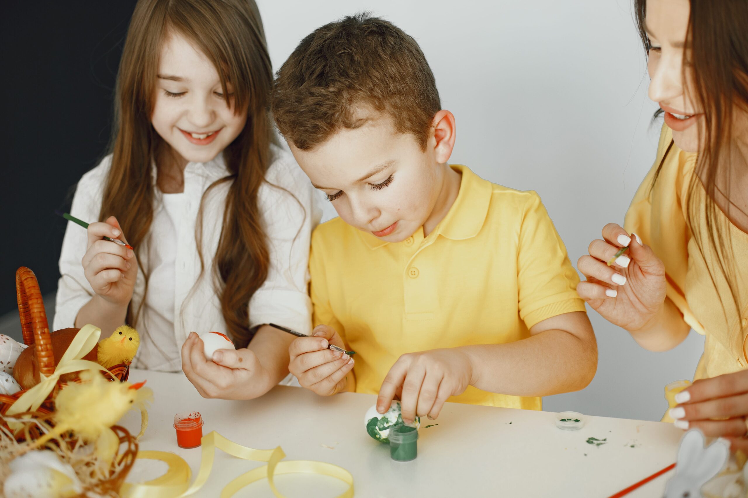 A young boy and girl, guided by an adult, engage in an egg-painting activity at a white table. The children focus intently, practicing fine motor skills and creativity—essential life skills for children with autism. The adult gently supports their learning, highlighting the value of structured, hands-on activities in autism therapy. A basket with decorated eggs and yellow ribbons adds a festive, sensory-rich element to the scene.