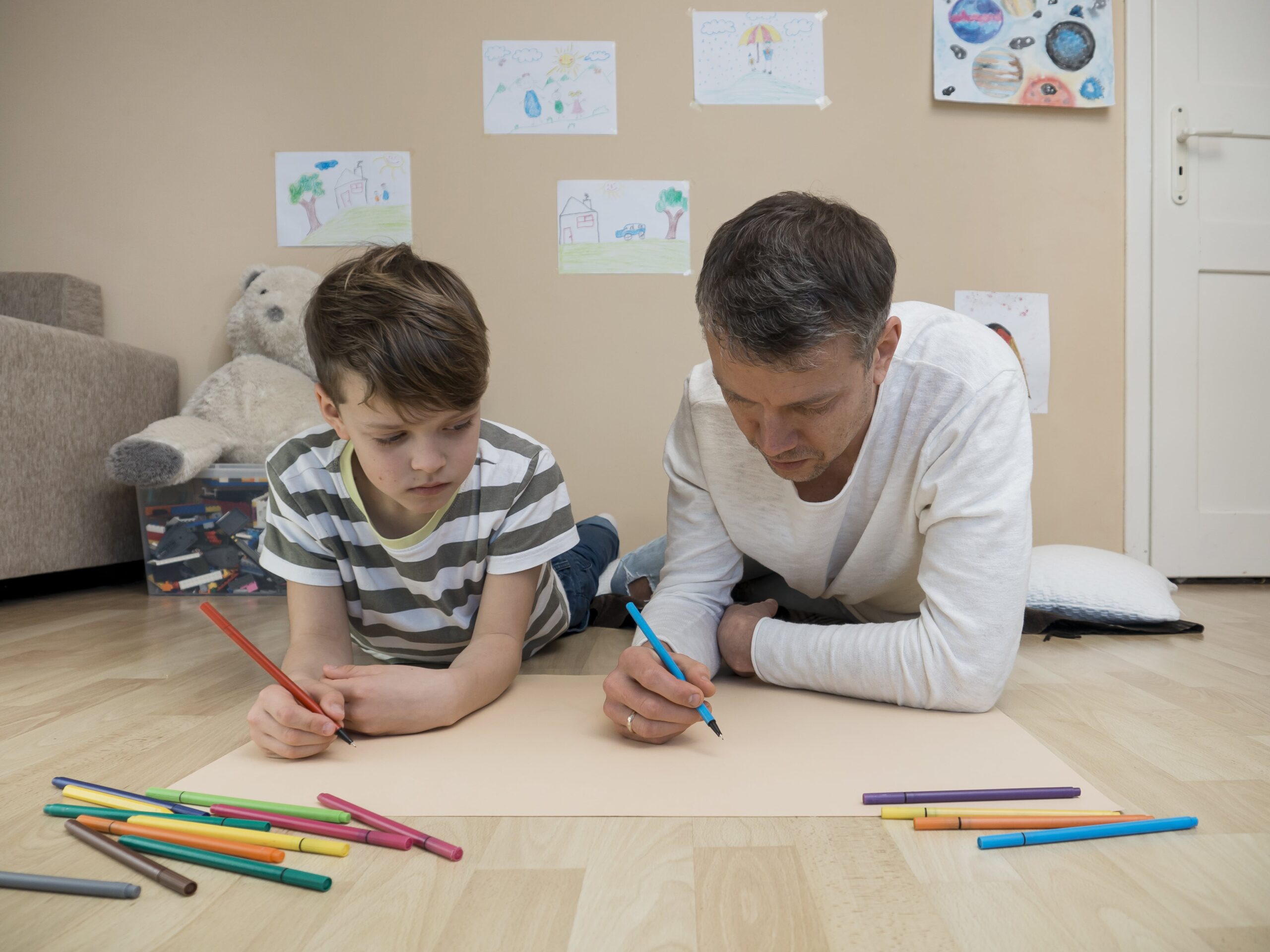 A father and son lie on the floor drawing together, building communication and focus—key life skills for children with autism—in a warm, creative environment.